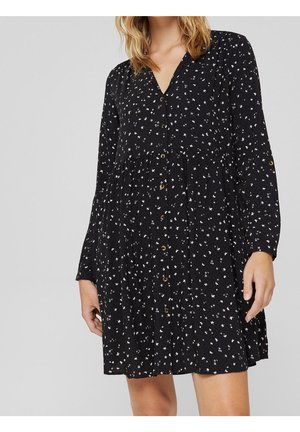 Femme portant une robe boutonnée noire à manches longues avec un petit motif floral blanc, coupe ample et col en V.