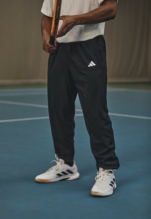 Svarta träningsbyxor med resårmidja och sidofickor, åtföljda av vita sneakers med svarta ränder. En tennisracket hålls i handen.