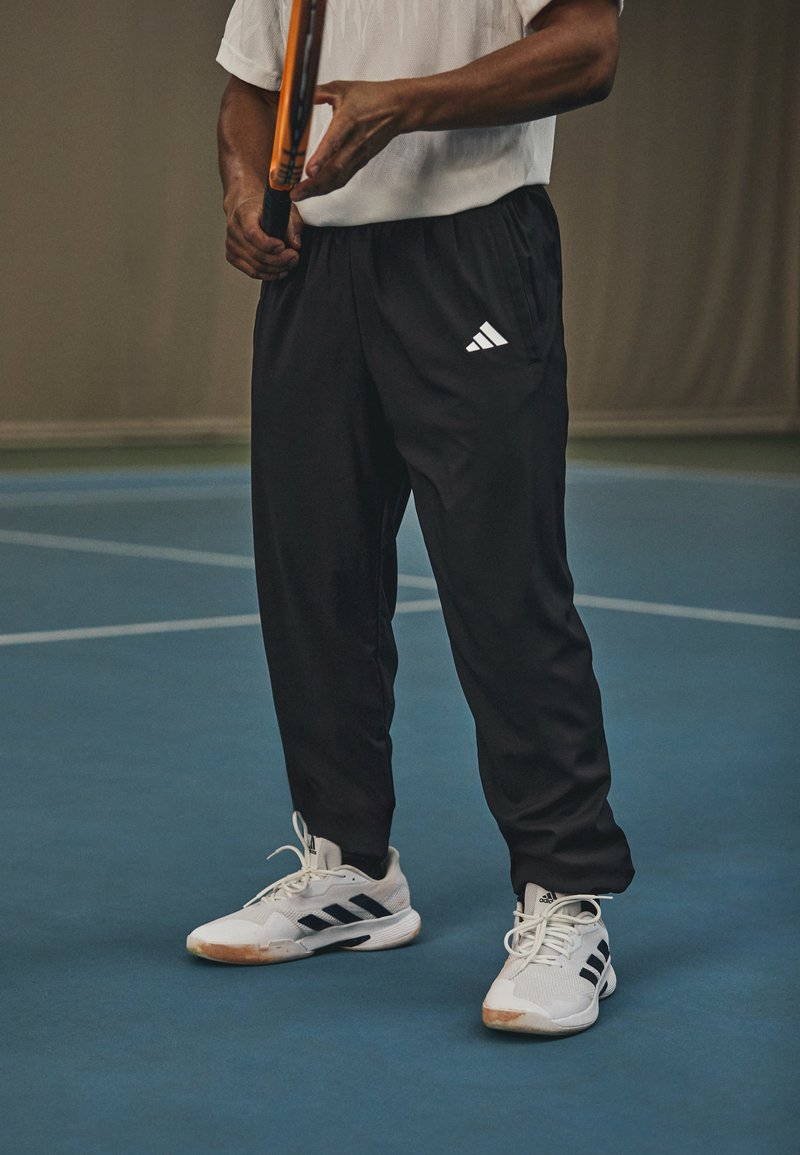 Un caleçon de sport noir avec une taille élastique et des poches latérales, associé à des baskets blanches avec des rayures noires. Une raquette de tennis est tenue à la main.