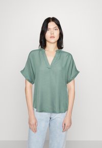 Grön kortärmad topp med V-ringning och avslappnad passform, tillverkad av ett mjukt material med en subtil lyster. Matchas med ljusblå jeans.