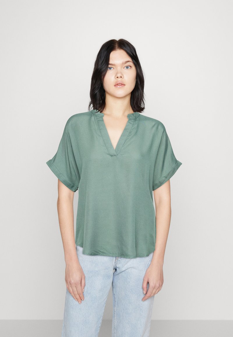 Grön kortärmad topp med V-ringning och avslappnad passform, tillverkad av ett mjukt material med en subtil lyster. Matchas med ljusblå jeans.