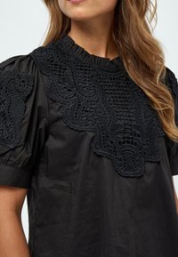 Blusa nera con un design in pizzo testurizzato sulla parte frontale, con collo arricciato e maniche corte a sbuffo. Finitura in tessuto liscia.