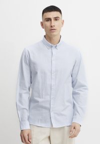 Camisa de botones a rayas azul claro y blanca, con mangas largas y cuello; presenta una textura suave y un diseño clásico.