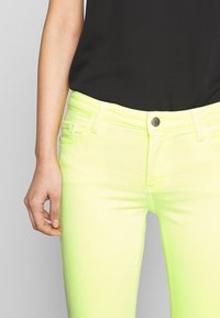 Jeans amarillos neón brillantes con un diseño ajustado, que cuentan con un estilo clásico de cinco bolsillos y un cierre de botón. Textura de tela suave.