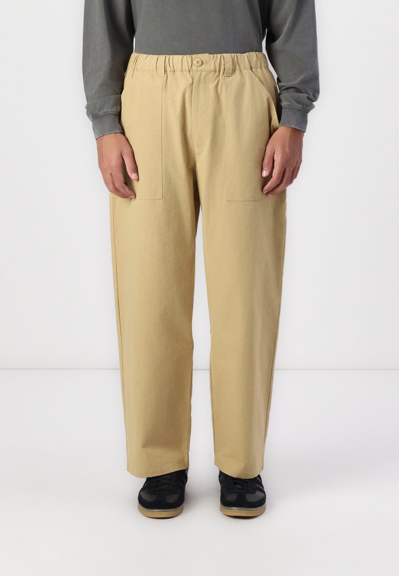 Beige, elastic waistband pants with two frontal pockets, loose fit, and cotton material, paired with black sneakers.