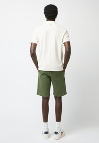Polo shirt crème, manches courtes, col avec détail de bouton ; associé à un short vert, coupe décontractée, et des baskets blanches avec des accents colorés.