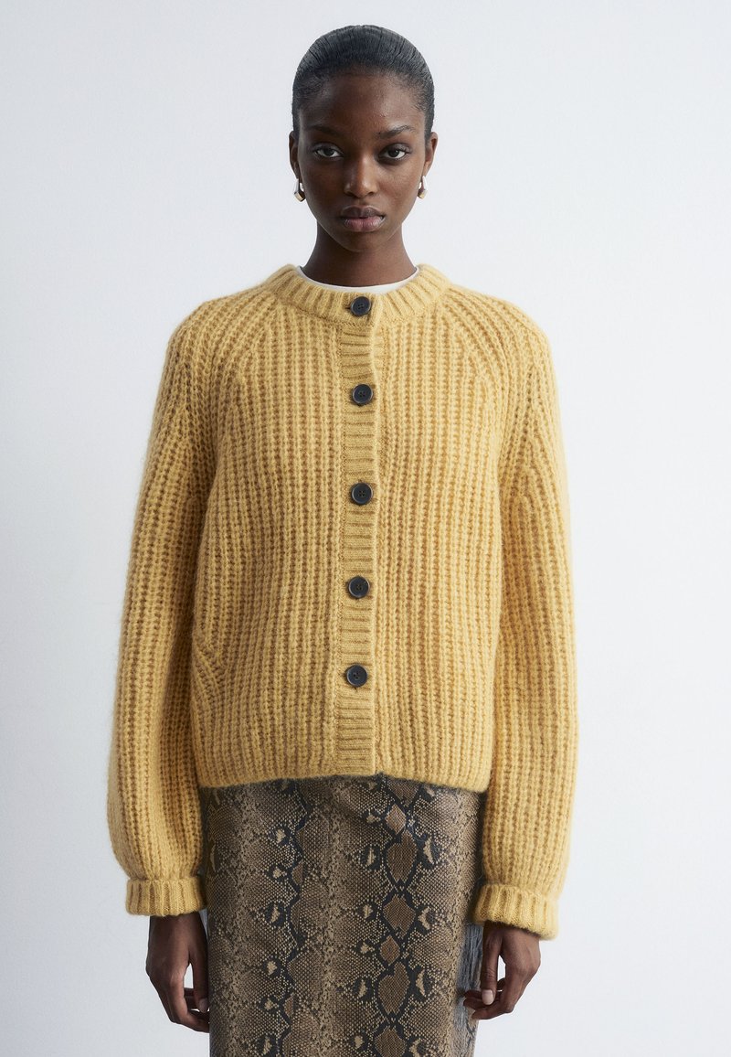 Yellow knitted cardigan with a ribbed texture, featuring a round neckline, black buttons, and long sleeves. Paired with a snakeskin print skirt.