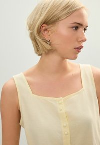 Top jaune à encolure carrée avec cinq boutons blancs sur le devant. Tissu en coton doux, larges bretelles et design léger.
