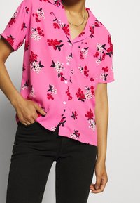 Kortärmad rosa skjorta med blommönster i rött, vitt och svart, med en ficka och knäppning fram, tillsammans med svarta byxor.