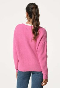 Overmaat roze gebreid sweater met een gestructureerd ribbelpatroon, ronde halslijn en lange mouwen. Gecombineerd met blauwe spijkerbroek.