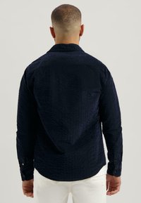 Man met kort haar, gekleed in een textuur donkerblauw overhemd met lange mouwen en witte broek, gezien van achteren tegen een effen achtergrond.