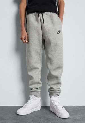 Personne portant un pantalon de survêtement Nike gris clair et des baskets montantes blanches Nike, debout devant un fond uni.