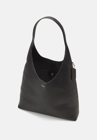 Sac à épaule en cuir noir Coach avec une large sangle simple, petit logo doré à l'avant, et étiquette noire attachée avec une chaîne dorée.