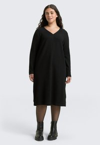Zwarte jurk met lange mouwen en V-hals, met een textuurstof en een losse pasvorm, gecombineerd met enkellaarsjes en sheer panty's.