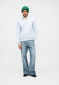 Homme portant un bonnet en laine vert, un sweat à demi-fermeture éclair bleu clair sur une chemise beige, un jean large bleu clair et des chaussures foncées, debout.