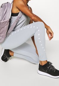 Leggings grigi chiari con branding bianco lungo il lato, abbinati a scarpe da ginnastica nere con tomaia testurizzata e suola bianca.