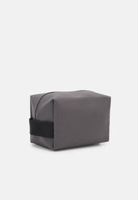Grå rektangulær kosmetikkveske laget av glatt, matt materiale. Har glidelås lukking og et svart stoffbånd på siden.