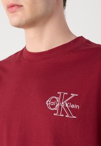 Bordo kokvilnas T-krekls ar apaļu kakla izgriezumu un baltu izšūtu Calvin Klein logo kreisajā krūtī. Īsas piedurknes, ikdienas piegriezums.