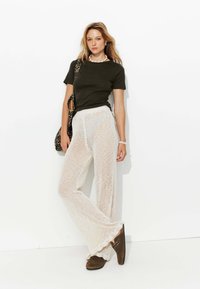 Pantalons larges en tricot blanc, t-shirt noir ajusté et chaussures en suède marron avec des bords festonnés. Les accessoires comprennent un sac à motif léopard et un collier.