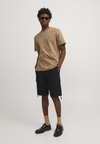 Brun kortærmet t-shirt, sorte cargo shorts med lommer, beige sokker og sorte sko. Modellen har solbriller på og står med hænderne i lommerne.