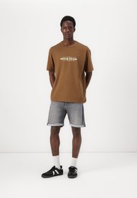 Brauni īsām piedurknēm T-krekls ar "Dockers" logo, apvienots ar gaiši denima šortiem un melniem sporta apaviem ar baltām svītrām. Iekļautas baltas zeķes.