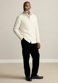 Cream cable-knit sweater with a half-zip collar, layered over a white shirt, paired with black corduroy pants and black loafers.
