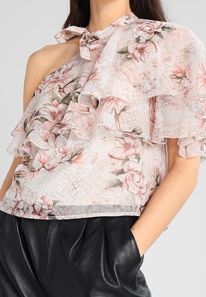 Frau trägt eine pinkfarbene, asymmetrisch gerüschte Bluse mit Blumenmuster und einer Schleife auf einer Schulter sowie schwarze Lederhosen, Hände in den Taschen.