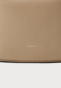 Bolsa de couro bege com acabamento texturizado, apresentando um logotipo em relevo dourado "FURLA" na parte da frente. Bordas suaves e design minimalista.