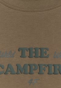 Gros plan d'un tissu en maille marron avec le texte en relief « THE CAMPFIR » en grandes lettres et des mots partiellement en cursive au-dessus et en dessous.