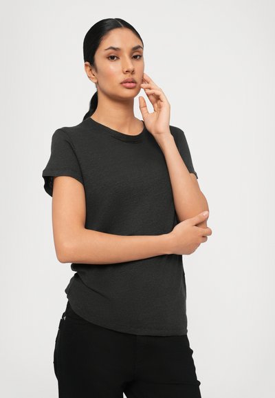 Mujer de cabello oscuro recogido, vestida con una camiseta negra ajustada de manga corta, posando con una mano cerca del rostro y la otra cruzada sobre su cuerpo.