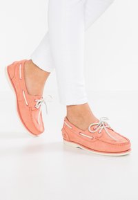 Kvinna som bär korallrosa båtklubbor med vita snören och vita byxor, med en fot något upphöjd.