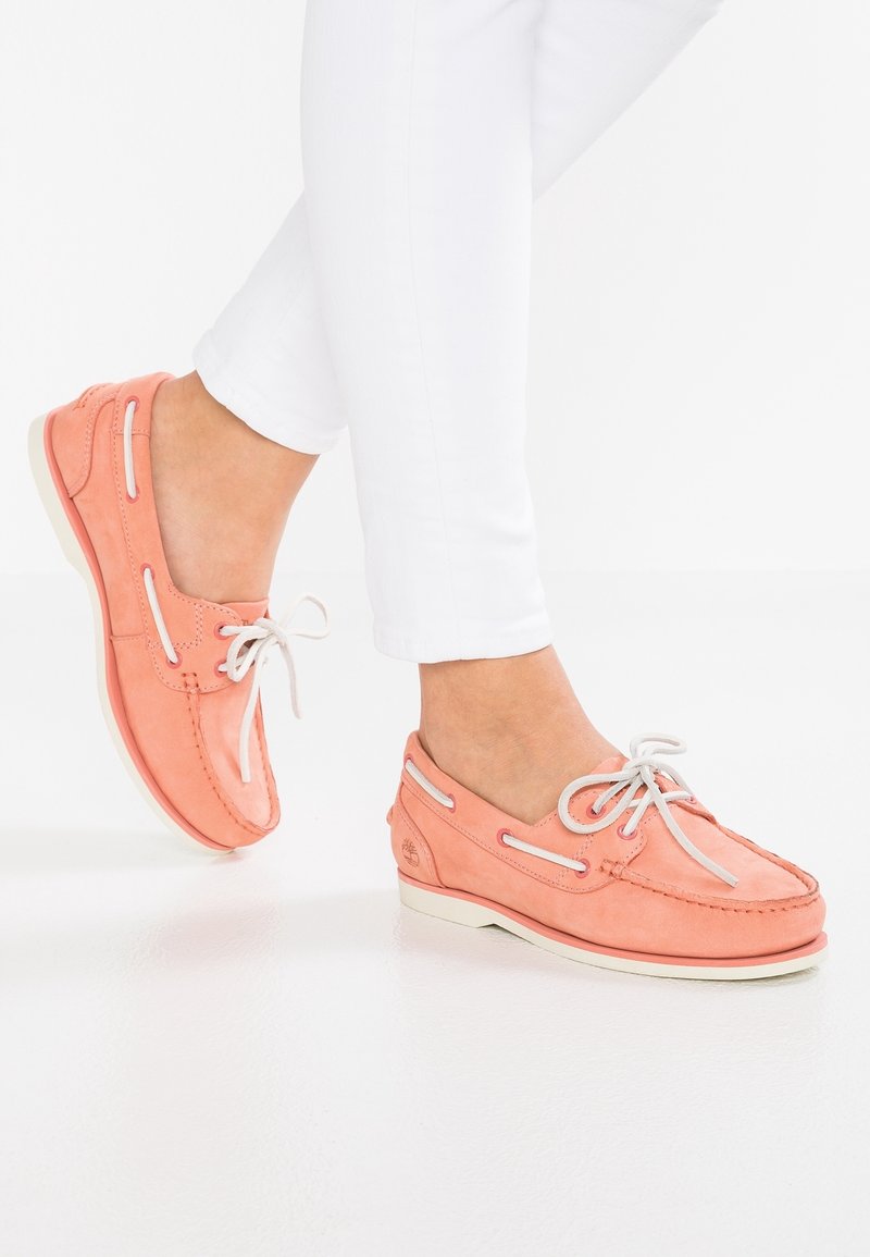 Kvinna som bär korallrosa båtklubbor med vita snören och vita byxor, med en fot något upphöjd.