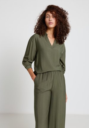 Frau mit lockigem Haar, die ein locker sitzendes olivgrünes Langarmshirt und passende weit geschnittene Hosen trägt, steht mit einer Hand in der Tasche.