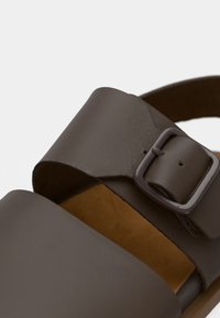 Καφέ δερμάτινο σανδάλι με φαρδιά λουρί και ρυθμιζόμενη πόρπη. Η σόλα έχει φυσικό χρώμα, με ομαλή υφή και μινιμαλιστικό σχεδιασμό.