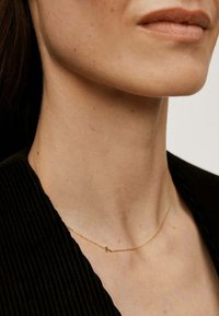 Collar de cadena dorada con un pequeño colgante de cruz. La cadena es delicada y tiene una textura suave, complementando una prenda negra y texturizada.