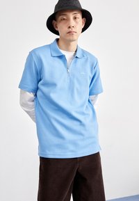 Polo azul com fecho em zíper e padrão texturizado, usado por cima de uma camisa de mangas longas branca, combinado com calças de veludo cotelê castanhas e um chapéu preto.
