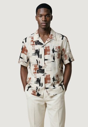 Mężczyzna ubrany w koszulę na guziki z krótkim rękawem, ozdobioną abstrakcyjnym wzorem czarnych, brązowych i beżowych pociągnięć pędzla, w połączeniu z jasnobeżowymi spodniami.