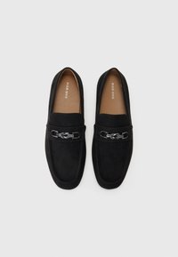 Czarne mokasyny zamszowe na płaskiej podeszwie, z ozdobnym wzorzystym srebrnym łańcuszkiem na przodzie. Gładka faktura i klasyczny design.