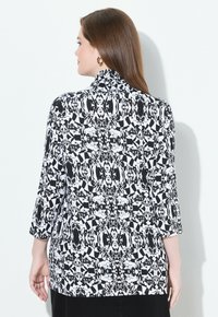 Schwarz-weiße gemusterte Bluse mit dreiviertel Ärmeln, ausgestattet mit abstrakten geometrischen Designs und einem hohen Kragen. Glatte Stoffoberfläche.