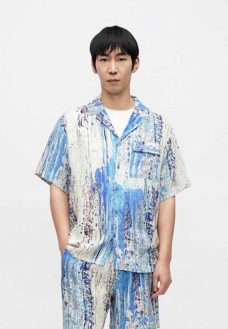 Camisa de manga corta y pantalones a juego en tejido ligero, con un patrón abstracto de gotas de pintura azul y blanca, y un bolsillo en el pecho izquierdo.