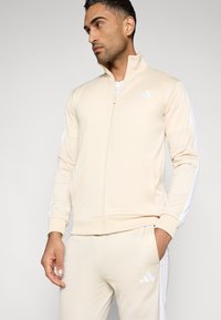 Μπεζ ζακέτα με φερμουάρ και υψηλό γιακά, κατασκευασμένη από λείο ύφασμα, με λευκές ρίγες κατά μήκος των μανικιών και λογότυπο στο στήθος.