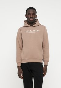 Felpa marrone in tessuto morbido con tasca a marsupio frontale. Presenta un cappuccio con lacci e logo bianco stampato sul petto. Pantaloni neri.