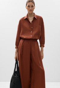 Camicia di lino color ruggine con bottoni, tasca e maniche lunghe, abbinata a pantaloni larghi coordinati; porta un borsa in tessuto nera.