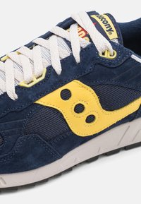 Navy blue sneaker with suede and mesh material, yellow accents, and three circular cutouts. Features white laces and a rubber sole.