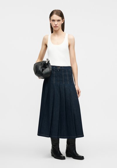 Γυναίκα που φοράει λευκό αμάνικο τοπ, σκούρα πλισέ τζιν φούστα, μαύρες μπότες και κρατάει μια μεγάλη μαύρη δερμάτινη τσάντα φάκελο.