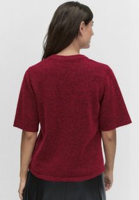 Czerwony sweter z krótkim rękawem wykonany z tkanej tkaniny, z okrągłym dekoltem i luźnym krojem. Materiał wydaje się miękki i ciepły.