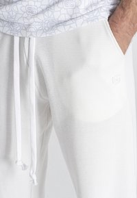 Joggers blancos texturizados con cintura elástica, cierre de cordón y bolsillo lateral. Presenta un sutil detalle de logo en la parte frontal.