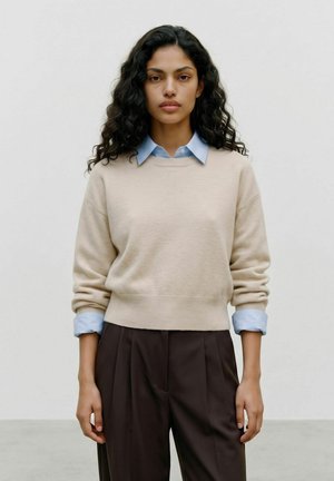 Kvinde med langt, krøllet mørkt hår iført en beige sweater over en lyseblå skjorte med krave og mørkebrune bukser, stående foran en ensfarvet baggrund.