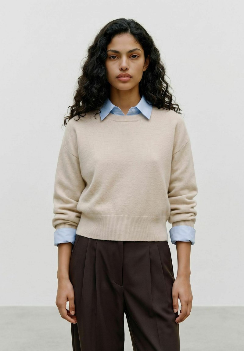Femme aux longs cheveux bouclés et foncés portant un pull beige par-dessus une chemise bleu clair à col et un pantalon marron foncé, debout devant un fond uni.