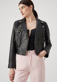 Giacca biker cropped in pelle nera con zip argentate, bottoni a pressione e tasche laterali, abbinata a pantaloni rosa chiaro.
